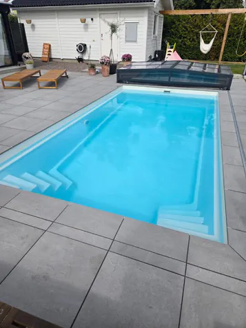 Glasfiberpool med trappa i båda ändar, grå plattor och öppet pooltak